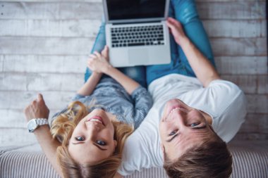 Bir dizüstü bilgisayar kullanarak rahat giysiler içinde güzel genç çift üst görünümü, kamera bakarak ve gri duvara karşı katta otururken gülümseyerek