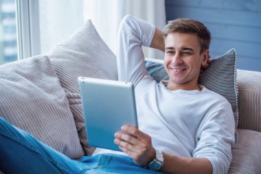 Rahat giysiler içinde Yakışıklı genç işadamı evde kanepede yatarken bir dijital tablet kullanıyor ve gülümsüyor