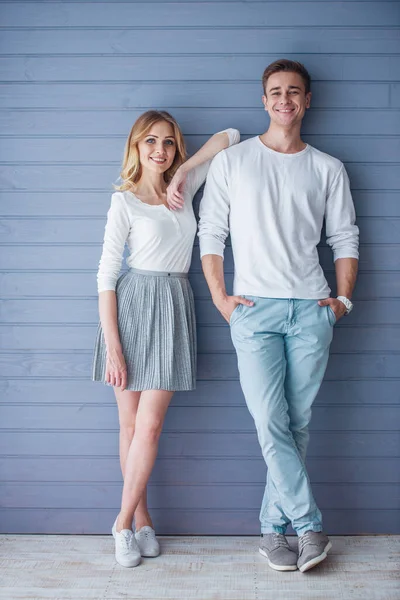 灰色の壁に立って カメラと笑顔を見てカジュアルな服を着た美しい若いカップルのフルレングスの肖像画 — ストック写真