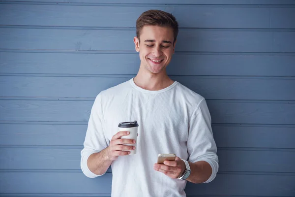 カジュアルな服を着たハンサムな若者は スマートフォンを使って 灰色の壁に立って コーヒーを飲んでいます — ストック写真