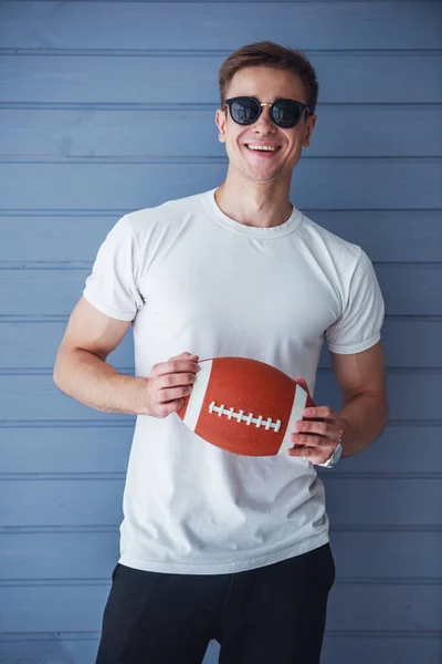 カジュアルな服とサングラスをかけたハンサムな若者がアメリカンフットボールのボールを持ち カメラを見て微笑み 灰色の壁に立っている — ストック写真