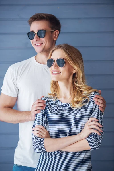 美丽的年轻夫妇在休闲的衣服和太阳眼镜的肖像 看着远方和微笑 站在灰色的墙壁 — 图库照片