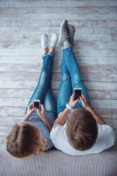 美丽的年轻夫妇在休闲服装的顶视图使用智能手机 而坐在地板上的灰色墙壁 — 图库照片
