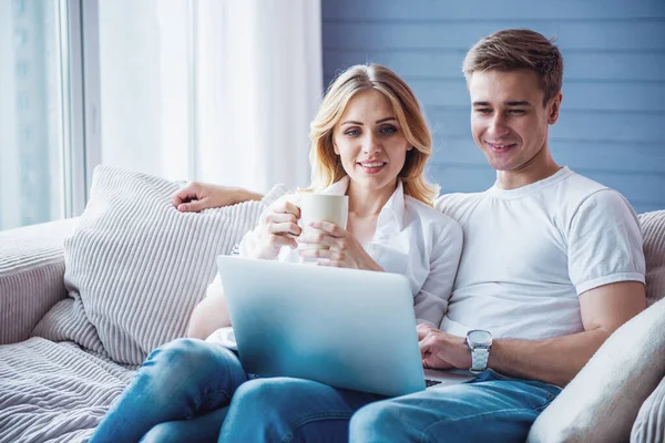 穿着休闲装的漂亮年轻夫妇正在使用笔记本电脑 看着相机 坐在家里的沙发上微笑 女孩拿着杯子 — 图库照片