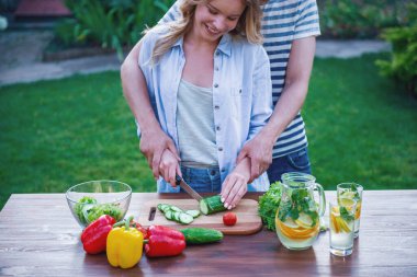 Genç çift kesme sebze salatası için görüntü açık havada yemek yaparken kırpılmış. Adam kızın arkasında duran ve ona yardım