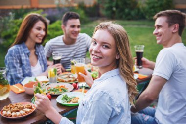 Sahip piknik açık havada genç insanlar. Güzel kız kameraya bakarak ve arkadaşları arka planda konuşurken gülümseyen