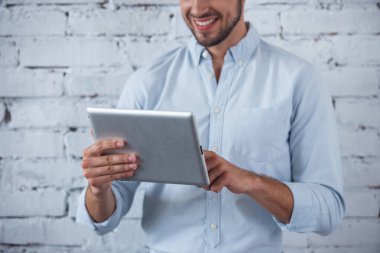 Bir dijital tablet kullanarak klasik gömlek yakışıklı genç işadamı kırpılmış görüntü ve gülümseyerek, beyaz tuğla duvar önünde duran