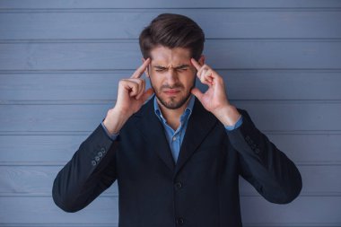 Klasik takım elbiseli yakışıklı genç yorgun işadamı acı içinde, yüzünü ellerini tutmak, gri duvarın önünde kapalı gözlerle ayakta