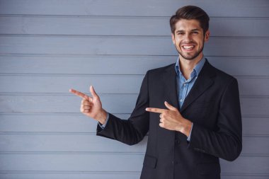 Klasik takım elbiseli yakışıklı genç başarılı işadamı yan işaret ediyor, kamera bakıyor ve gülümseyerek, gri duvarın önünde duruyor