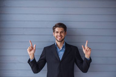 Klasik takım elbiseli yakışıklı genç başarılı işadamı yukarı yaslanıyor, kameraya bakıyor ve gülümsüyor, gri duvarın önünde duruyor