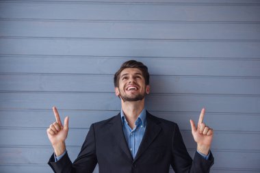 Klasik takım elbiseyakışıklı genç başarılı işadamı gülümsüyor, işaret ve yukarı bakıyor, gri duvarın önünde duruyor