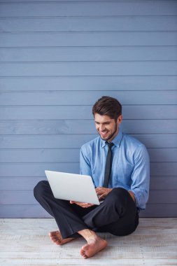 Bir dizüstü bilgisayar kullanarak klasik takım elbise yakışıklı genç başarılı işadamı Portresi ve gülümseyerek, gri duvarın önünde katta yalınayak oturan