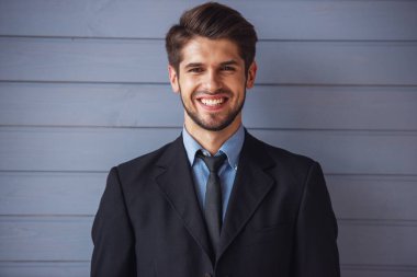 Klasik takım elbiseli yakışıklı genç başarılı işadamıportresi kameraya bakarak ve gülümseyerek, gri duvarın önünde duruyor