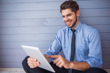 Klasik takım elbiseli yakışıklı genç işadamı bir dijital tablet kullanıyor ve gülümseyerek, gri duvarın önünde oturuyor