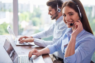 Yan görünümü ofiste çalışırken dizüstü bilgisayarlar kullanarak kulaklık yakışıklı işadamı ve güzel genç iş kadını. Kız kameraya bakıyor