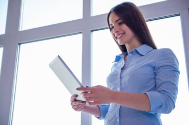 Güzel genç iş kadını dijital tablet kullanma ve gülümseyerek, önce pencere ayakta klasik gömlek düşük açılı görünümü
