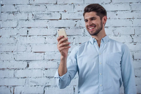 Klasik gömlek yakışıklı genç işadamı beyaz tuğla duvar önünde duran, bir akıllı telefon kullanıyor ve gülümseyerek