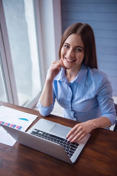 Güzel genç iş kadını bir dizüstü bilgisayar kullanarak, kameraya bakıyor ve ofiste çalışırken gülümseyen
