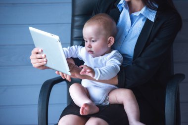 Güzel iş kadını Klasik takım elbise ve onun tatlı küçük bebek dijital tablet kullanma ve birlikte, vakit ofis koltuğu gri arka plan üzerinde oturan kırpılmış ederken gülümseyen