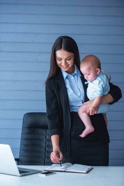 Klasik takım elbisegüzel iş bayan kollarında onu sevimli küçük bebek tutuyor, notlar yapma ve ofiste çalışırken gülümseyerek