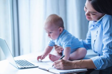 Klasik takım elbisegüzel iş bayan cep telefonu konuşuyor, notlar yapma ve gülümseyerek, onun sevimli küçük bebek masada otururken ofiste çalışıyor