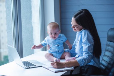 Klasik takım elbisegüzel iş bayan cep telefonu konuşuyor, notlar yapma ve gülümseyerek, onun sevimli küçük bebek masada otururken ofiste çalışıyor
