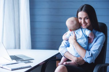 Sevimli küçük bebeği kollarında holding, kameraya bakıyor ve ofiste otururken gülümseyen güzel iş kadını