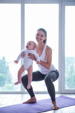 Spor giyim güzel genç anne büyüleyici küçük bebeği pencere karşı bir mat ile yoga yaparken gülümsüyor