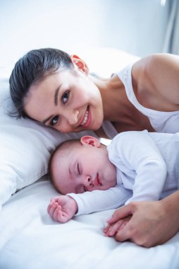 Kameraya bakarak ve ona şirin bebek evde yatakta uyurken gülümseyen güzel genç anne yan görünüm