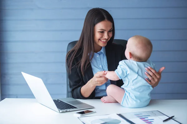 Beautiful business lady in classic suit is smiling while playing with her cute little baby in office