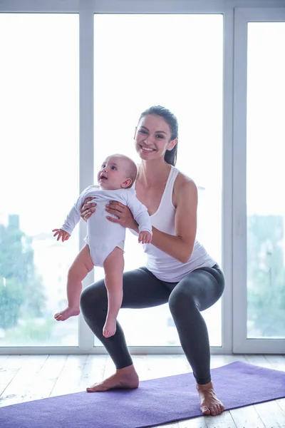 Beautiful young mom in sports wear is smiling while doing yoga with her charming little baby on a mat against window