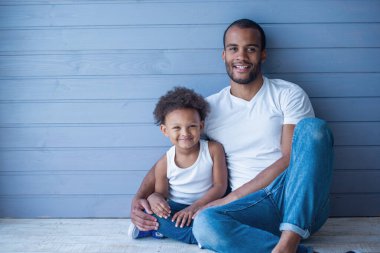 Yakışıklı genç Afro Amerikalı baba ve onun sevimli çocuk portre kamera bakarak ve gülümseyerek, gri duvar önünde oturan