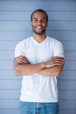 Rahat giysiler içinde Yakışıklı genç Afro Amerikalı adam kamera bakıyor ve gülümseyerek, gri arka plan üzerinde çapraz kolları ile ayakta