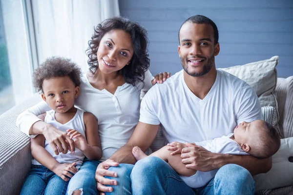 Portrait of beautiful young Afro American parents and their cute children looking at camera and smiling while sitting on sofa at home. Little baby is sleeping in dad\'s arms