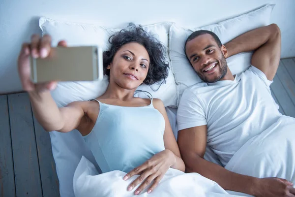 Beautiful young Afro American couple is making selfie using a smart phone and smiling while lying in bed at home
