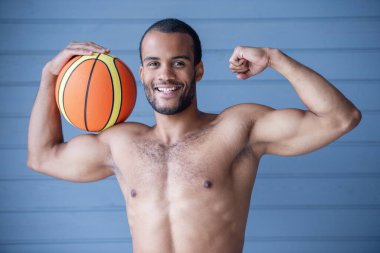 Basketbol topu tutan yakışıklı çıplak sporcunun portresi, kaslarını gösteriyor, kameraya bakıyor ve gülümsüyor, gri arka planda.