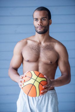 Gri şortlu, kendine güvenen yakışıklı bir sporcunun portresi. Elinde gri arka planda basket topu var.