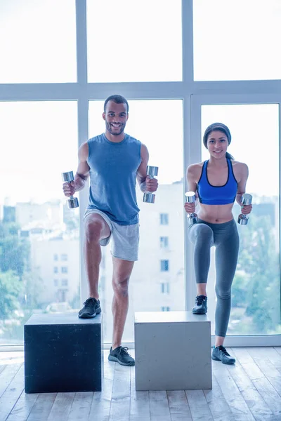 stock image Beautiful couple in sports clothes is working out with dumbbells and wooden boxes, looking at camera and smiling