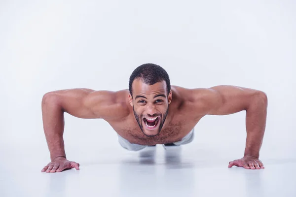 Handsome sportsman is doing push-ups, looking at camera and screaming while working out topless, isolated on white background