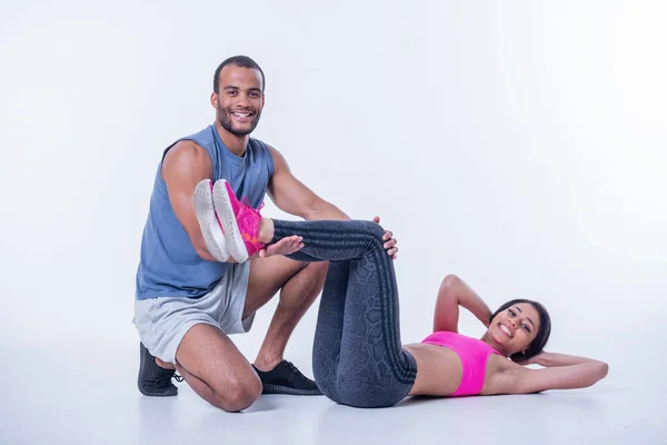 stock image Beautiful sportswoman is doing abs, handsome Afro American trainer is holding her legs. Both are looking at camera and smiling, isolated on white background