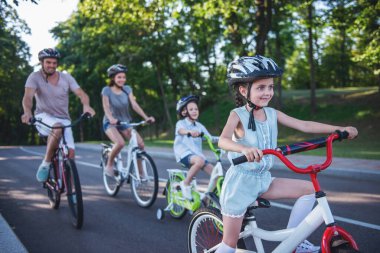 Mutlu aile açık havada bisiklet sürme ve gülümseyerek. Ön planda küçük kız