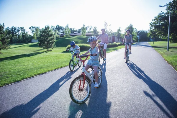 Happy family is riding bikes outdoors and smiling. Little girl in the foreground is looking at camera