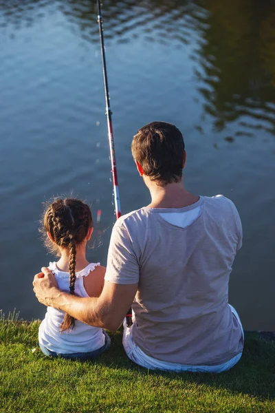 Back view of handsome father and his beautiful little daughter catching fish in the pond using a fishing rod, sitting on the ground