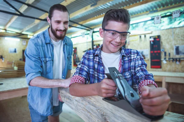 Handsome teenage carpenter in protective glasses and his father are smiling while working with wood and carpenter's plane in the workshop