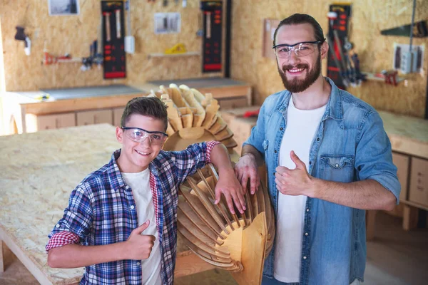 Handsome teenage carpenter and his father in protective glasses are showing Ok sign, looking at camera and smiling while standing near their done wooden object in the workshop