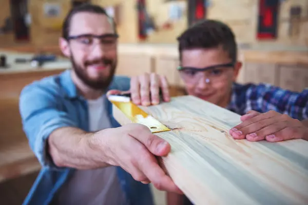Handsome teenage carpenter and his father in protective glasses are measuring wood and smiling while working in the workshop