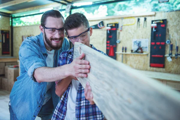 Handsome teenage carpenter and his father in protective glasses are examining wood and smiling while working in the workshop