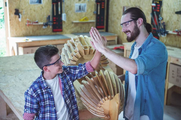 Handsome teenage carpenter and his father in protective glasses are giving high five and smiling while standing near their done wooden object in the workshop