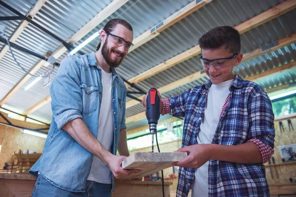 Handsome teenage carpenter and his father in protective glasses are smiling while working with wood and hammer drill in the workshop