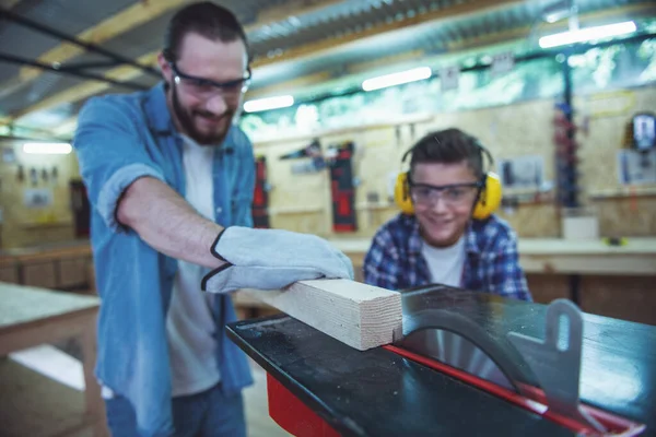 Handsome teenage carpenter and his father in protective glasses are smiling while cutting wood using a circular saw table in the workshop